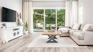 A modern living room with a flat-screen TV, white furniture, a wooden coffee table, and a large sliding glass door leading to an outdoor area.
