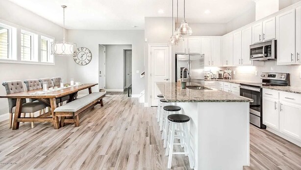 A modern kitchen with white cabinetry, a central island with bar stools, a dining area with a wooden table and chairs, and a large wall clock.