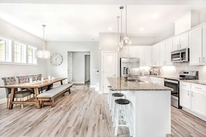 Een moderne keuken met witte kastjes, een centraal eiland met barkrukken, een eethoek met een houten tafel en stoelen, en een grote wandklok.