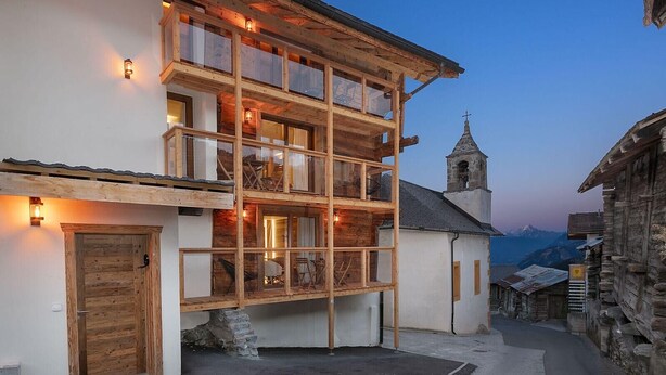 A multi-story wooden building with balconies, a stone staircase, and a church in the background. - Pilànous Residence - Junior Suite - Swiss Alps (Haute-Nendaz)