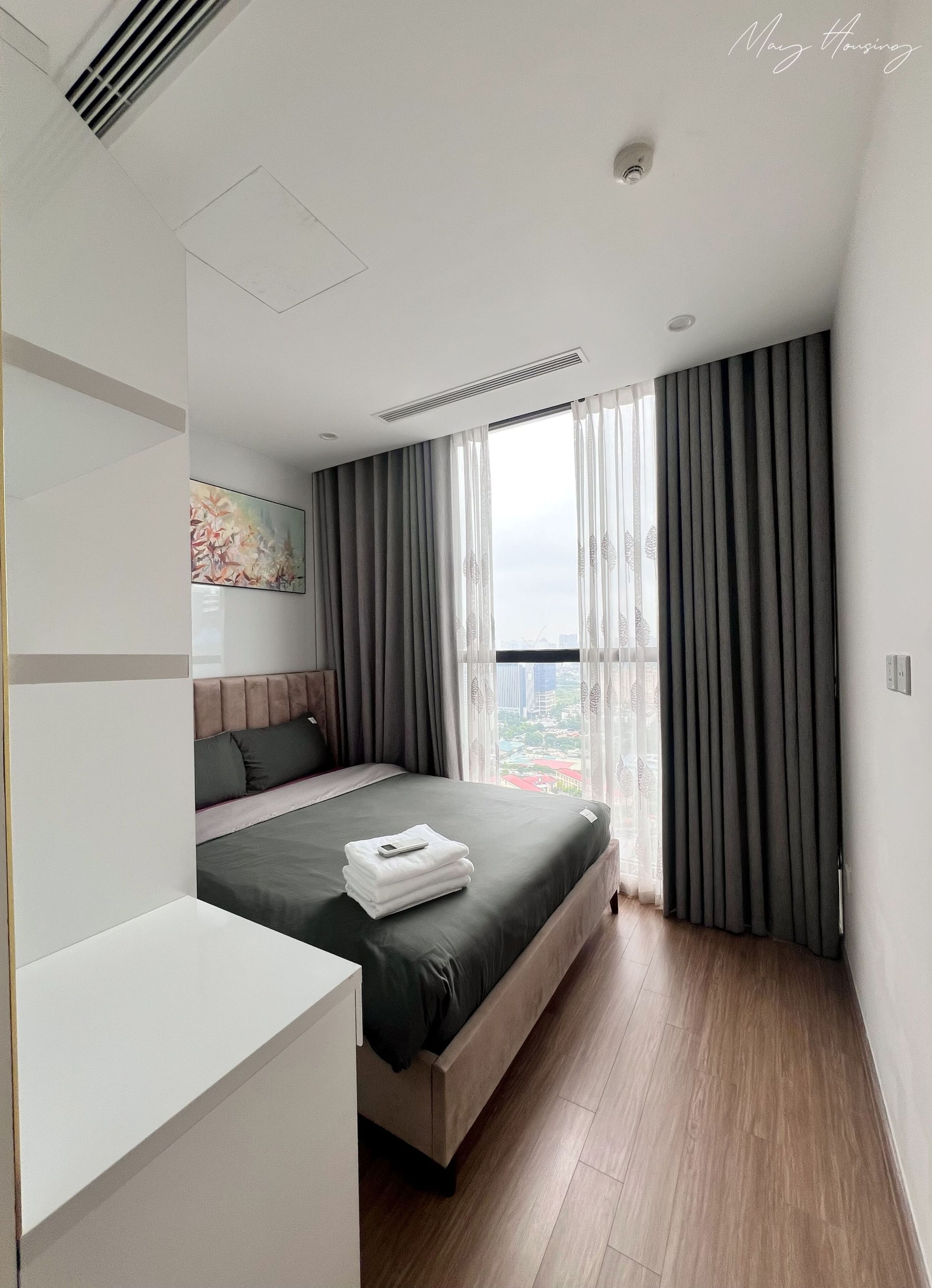 A modern hotel room with a large bed, a bedside table, and a view of the city through sheer curtains.