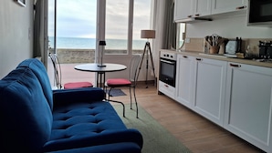 A modern kitchen with white cabinets, a microwave, and a stove. There is a dining area with a table and chairs, and a large window offering a view of the sea. - Nouveaute : Front DE MER Neuf Bien Équipé Belle VUE MER Pour Sejour Parfait (AGON COUTAINVILLE)