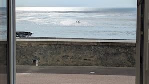 A view through a window of a beach with a stone wall and a paved walkway. - Nouveaute : Front DE MER Neuf Bien Équipé Belle VUE MER Pour Sejour Parfait (AGON COUTAINVILLE)