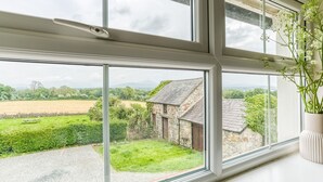 Vista desde una ventana a un edificio de piedra con tejado inclinado, rodeado de vegetación y campos.