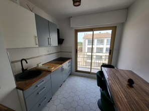Cocina moderna con un lavamanos, encimera de madera y armarios grises. Se ve una mesa de comedor con sillas, y un balcón con vistas a edificios a través de una ventana.