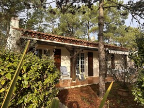 A rustic house with a tiled roof, a porch with chairs, and a garden with pine trees.