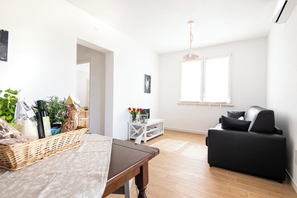 Ein modernes Wohnzimmer mit Holzboden, einer schwarzen Couch, einem weißen Beistelltisch mit Blumen in einer Vase und einem Korbkörbchen auf einem Tisch.
