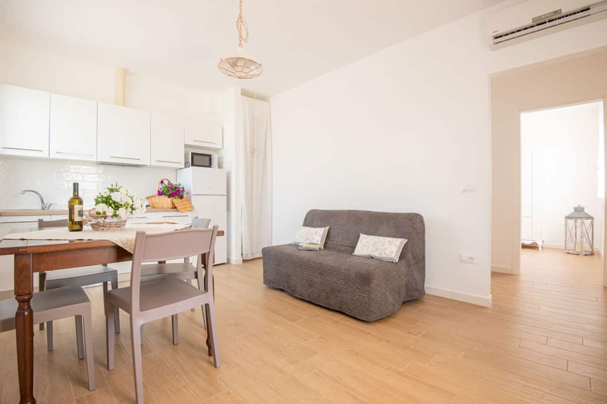 Eine moderne Küche mit weißen Schränken, einem Esstisch mit Stühlen und einem grauen Sofa in einem hellen Wohnbereich mit Holzboden.