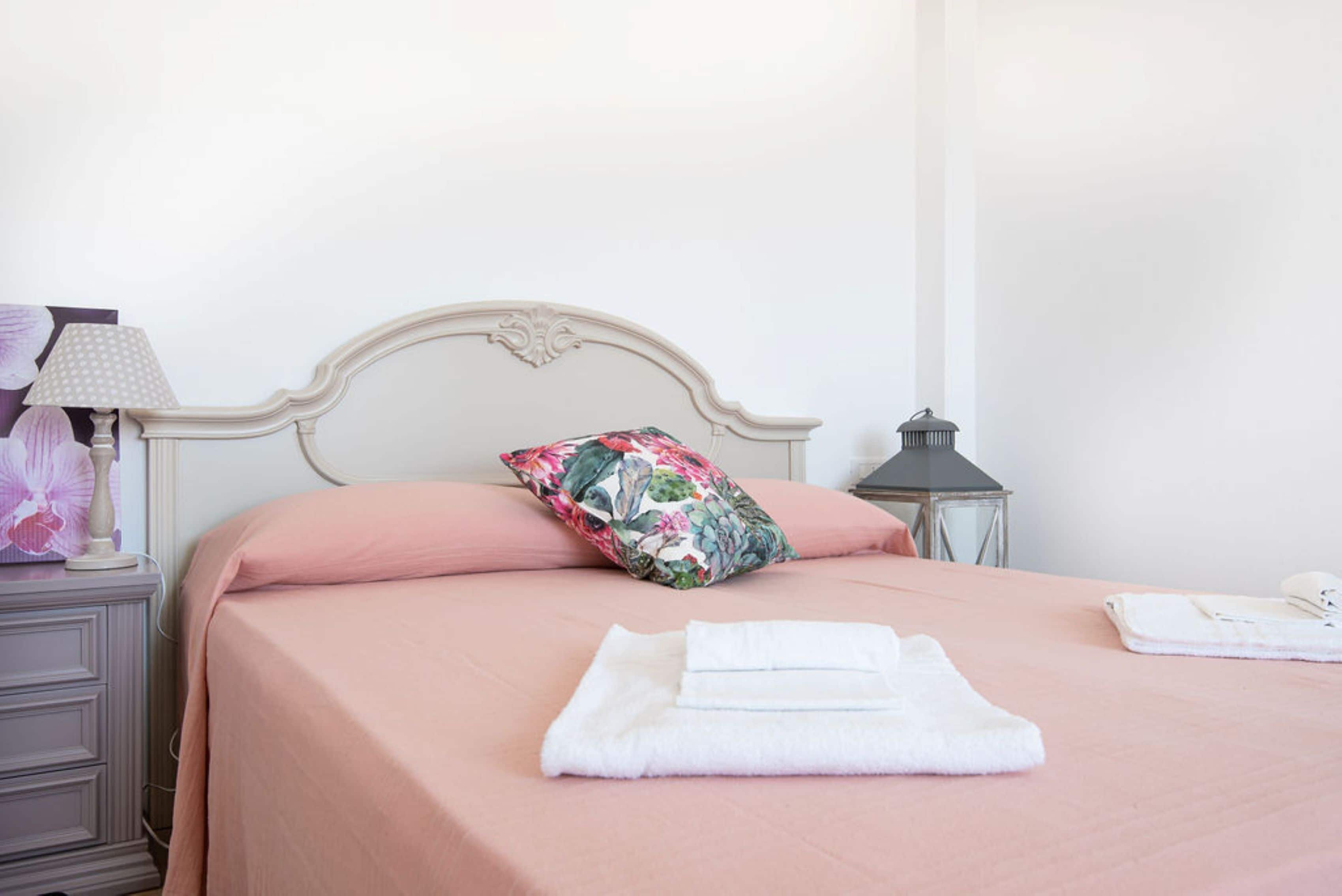 Ein ordentlich bezogenes Bett mit einer rosa Tagesdecke, einem blumigen Kopfkissen und einem weißen Frotteehandtuch darauf.