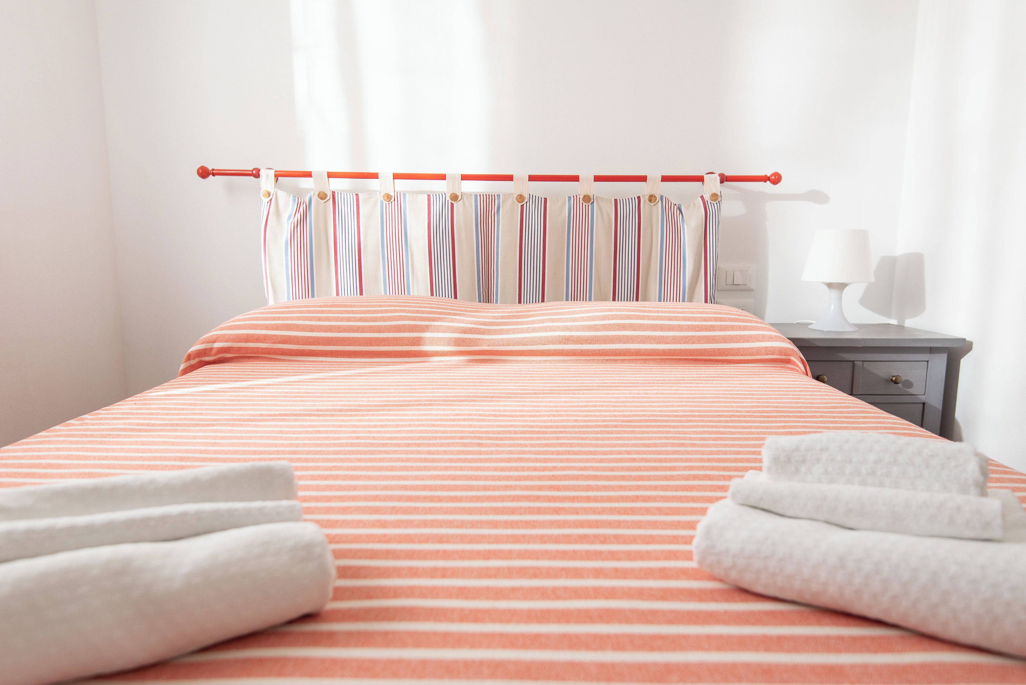 Ein ordentlich bezogenes Bett mit gestreifter Bettwäsche, ein rot-weiß gestreifter Vorhang und zwei gefaltete Handtücher auf dem Bett.