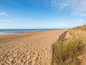 Ein weiter Sandstrand mit sanften Wellen, blauem Himmel und Dünen mit spärlicher Vegetation.
