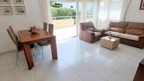 Una sala de estar con un sofá marrón, una mesa de centro, una mesa de comedor con sillas y un balcón con vistas a la vegetación.