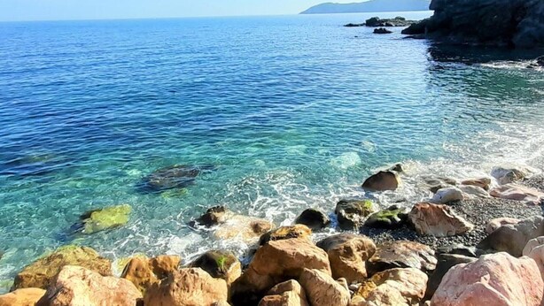 Escena costera con agua azul cristalina, costa rocosa y formaciones terrestres a lo lejos.