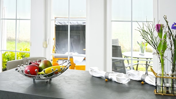 A dining area with a metal fruit bowl on a table, a vase with flowers, and a view of a balcony with a table and chairs.
