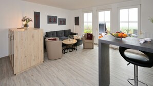 A modern living room with a grey sofa, a round coffee table, and a wooden cabinet. There are framed artworks on the wall and a window with a view of greenery.
