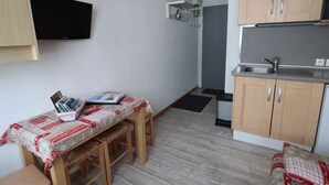 Une cuisine compacte avec des meubles en bois, un four à micro-ondes encastré et un évier. On y trouve un petit coin repas avec une table et des chaises, ainsi qu’un téléviseur fixé au mur.