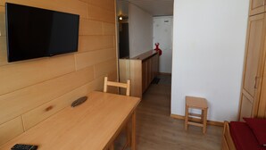 Une pièce avec une table en bois, un téléviseur fixé au mur, un petit tabouret et un lit avec un oreiller rouge.