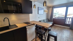 A compact kitchen with a microwave, sink, and a built-in dishwasher. There is a wooden dining table with two stools and a built-in bench with cushions. The room has a large window with a view of the city. - Comfort studio 4 pax at the foot of slopes with balcony (Tignes)