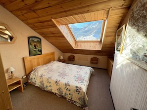 A bedroom with a sloped ceiling, a skylight, a bed with floral bedding, a wooden headboard, a nightstand, a framed picture, and a stone wall.