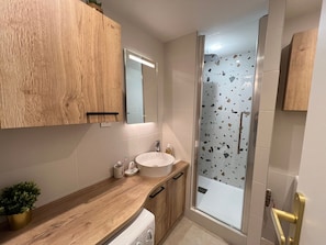 A modern bathroom with a wooden vanity, a white sink, a glass shower enclosure, and a small potted plant.