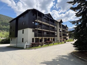 A multi-story building with balconies, a parking area, and a mountain backdrop.