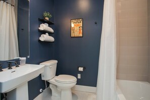 Un baño con lavamanos, inodoro y bañera blancos. Hay un perchero con toallas y un cuadro enmarcado en la pared.
