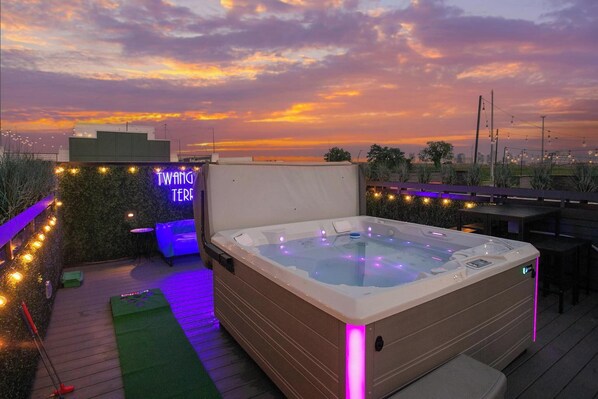 Un jacuzzi en una terraza en la azotea con un letrero de neón, rodeado de guirnaldas de luces y vistas del atardecer.