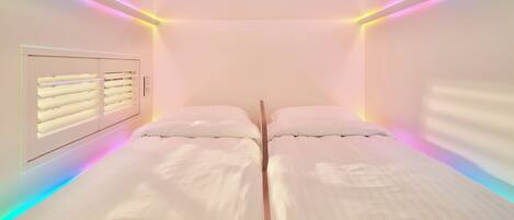A room with two beds, a window with blinds, and LED lighting along the ceiling.