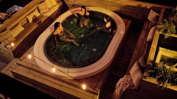 Un jacuzzi con dos personas, rodeado de una cubierta de madera y plantas.