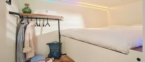 Habitación de hotel pequeña con una cama litera, un estante con ropa doblada y una maleta, y un perchero en la pared.
