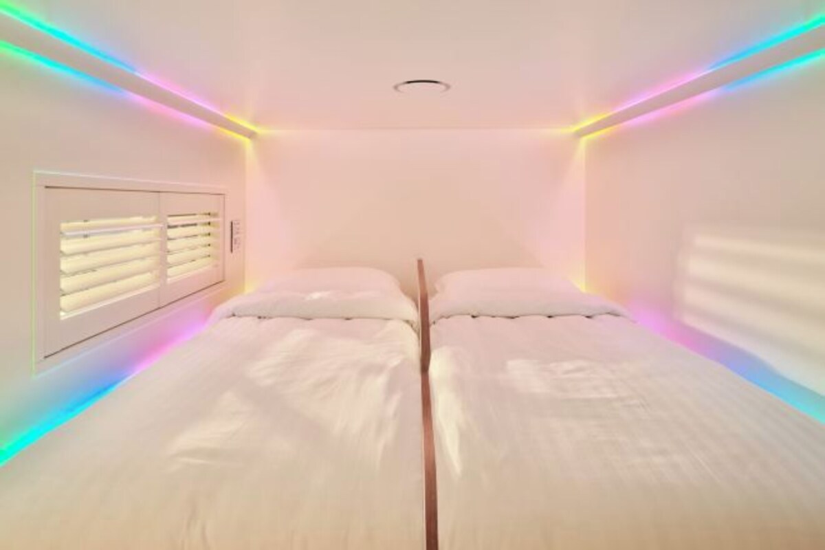 A room with two beds, a window with blinds, and colorful LED lighting along the ceiling.