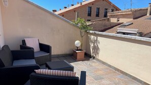Una terraza en la azotea con mobiliario de mimbre, una planta en maceta y vistas a edificios cercanos.