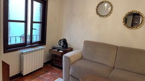 Salón con sofá color beige, un radiador, una ventana con vistas, una mesita de madera pequeña con una tetera y vasos, y dos espejos colgados en la pared.