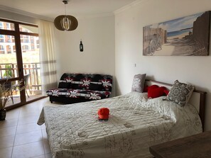 A bedroom with a bed, a sofa, a painting, and a view of buildings outside. - Brand New Studio 150 m to Sea  (Sveti Vlas)