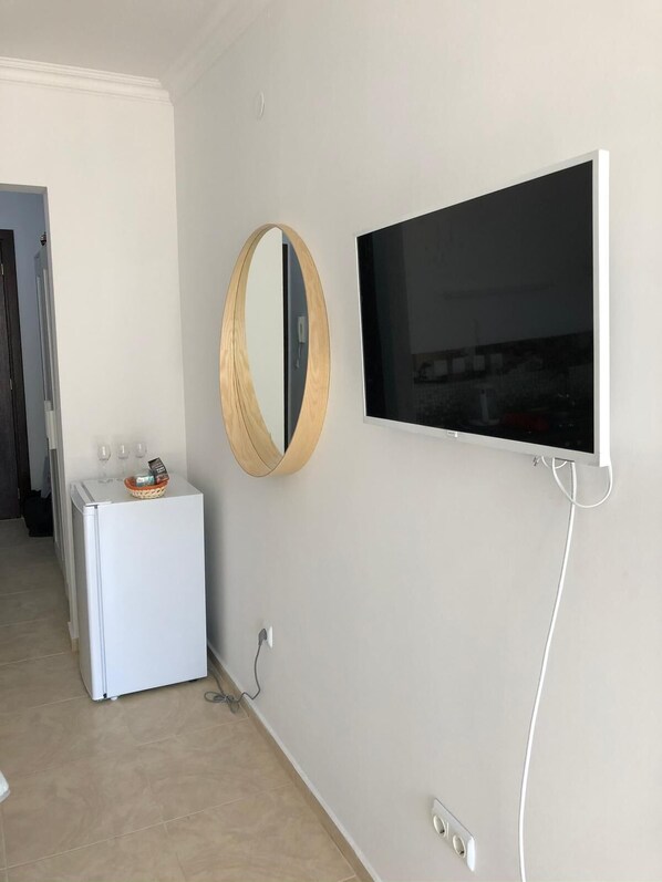 A modern living room with a flat-screen TV mounted on the wall, a wooden-framed mirror, and a white mini-fridge. - Brand New Studio 150 m to Sea  (Sveti Vlas)
