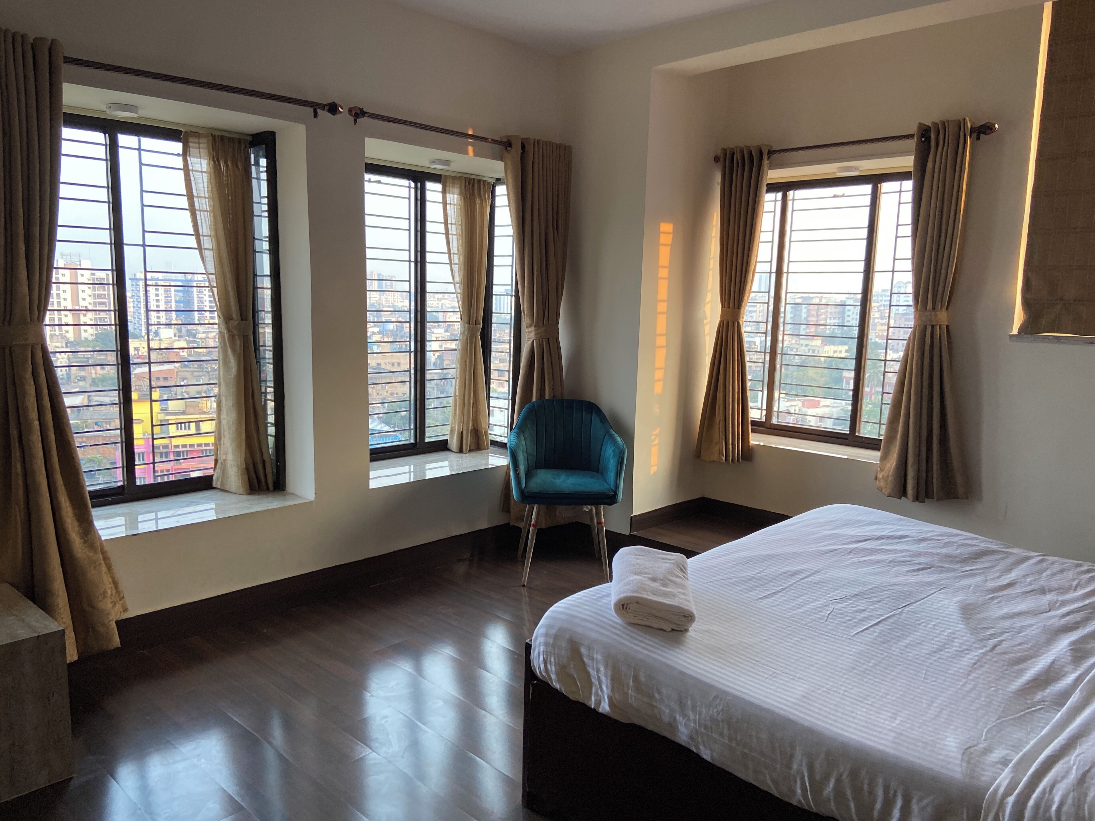 Habitación de hotel con una cama grande, una silla azul y varias ventanas con cortinas.