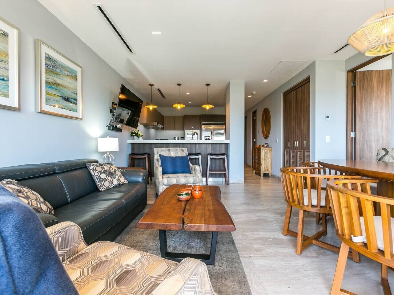 A modern living room with a sofa, a wooden dining table, and chairs. There is a kitchen area in the background with a countertop and cabinets.
