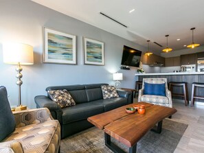 A modern living room with a sofa, a coffee table, and a patterned armchair. There is a kitchen area with a bar and a TV mounted on the wall.