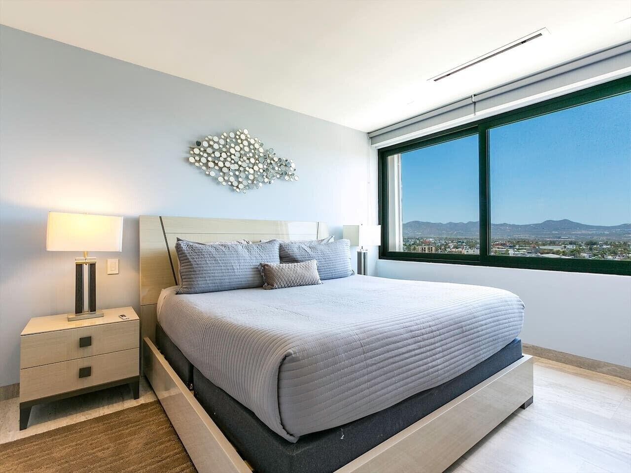 Ein modernes Schlafzimmer mit einem großen Bett, einem Nachttisch, einer Wandleuchte und Blick auf die Stadt durch ein großes Fenster.
