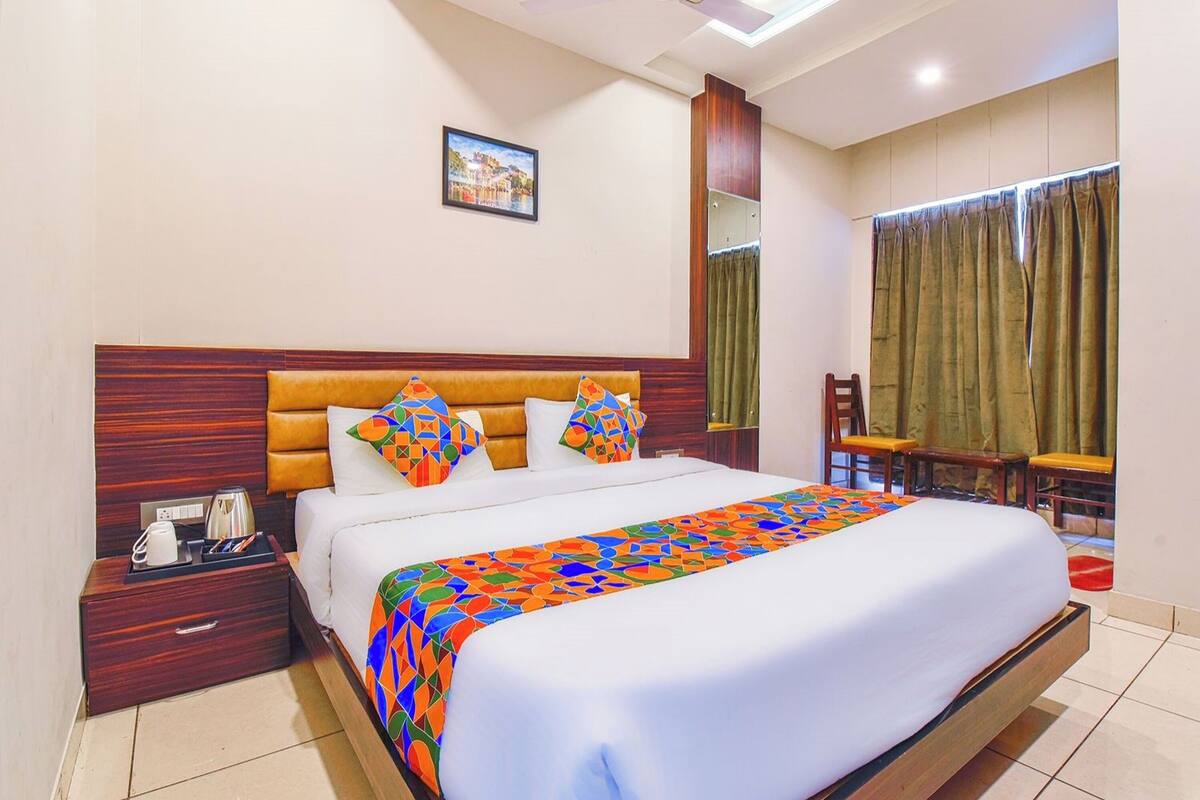 A hotel room with a large bed, a wooden headboard, a nightstand with a phone and a kettle, a window with curtains, and a small table with a chair.