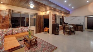 A modern living room with a sofa, dining table, and chairs. There is a fan, a refrigerator, and a large window. - Blossom Stay (Mahabaleshwar)