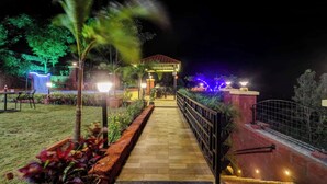 A nighttime view of a pathway leading to a building with a red roof, surrounded by greenery and illuminated by ambient lighting. - Blossom Stay (Mahabaleshwar)