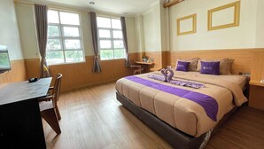 A hotel room with a large bed, a desk, and a chair. There are two windows with curtains, a wooden headboard, and a small table with a plant. - Take Guesthouse 1 (Jambi)