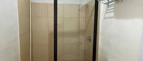 Un pequeño espacio de ducha cerrado con puerta de cristal, baldosas color beige y un inodoro blanco.