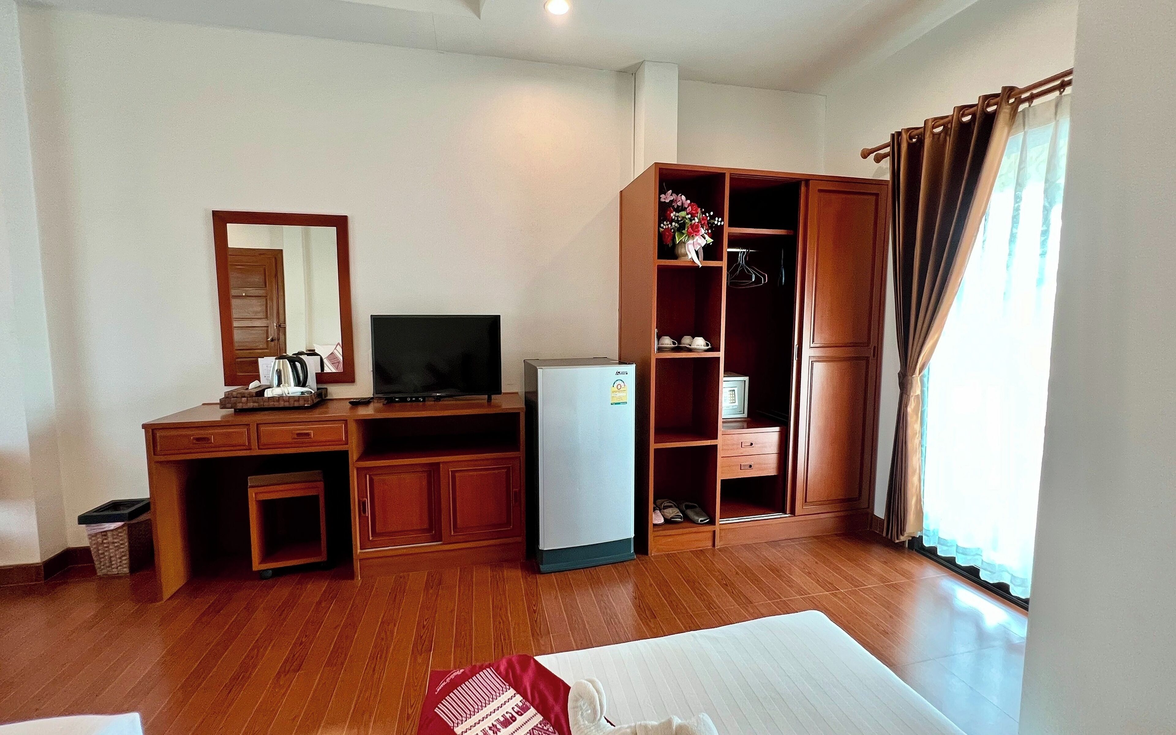 A hotel room with a wooden desk, a television, a refrigerator, a wardrobe, and a bed.