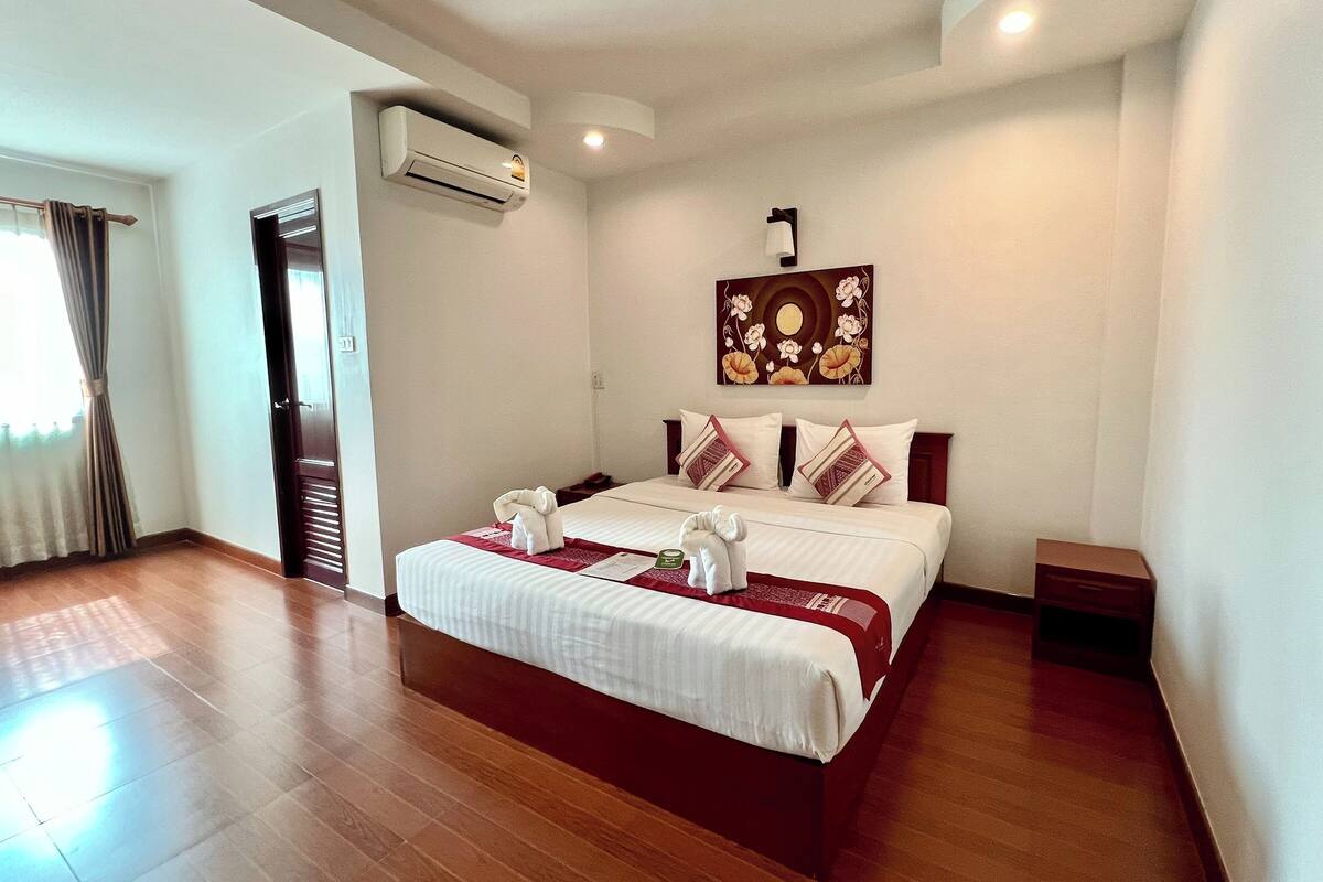A hotel room with a wooden floor, a bed with white bedding, a red and white throw, and an air conditioning unit on the wall.