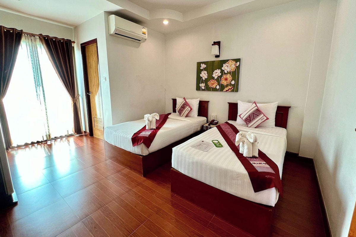 A hotel room with two beds, a painting on the wall, an air conditioning unit, and a view of the outside through the window.