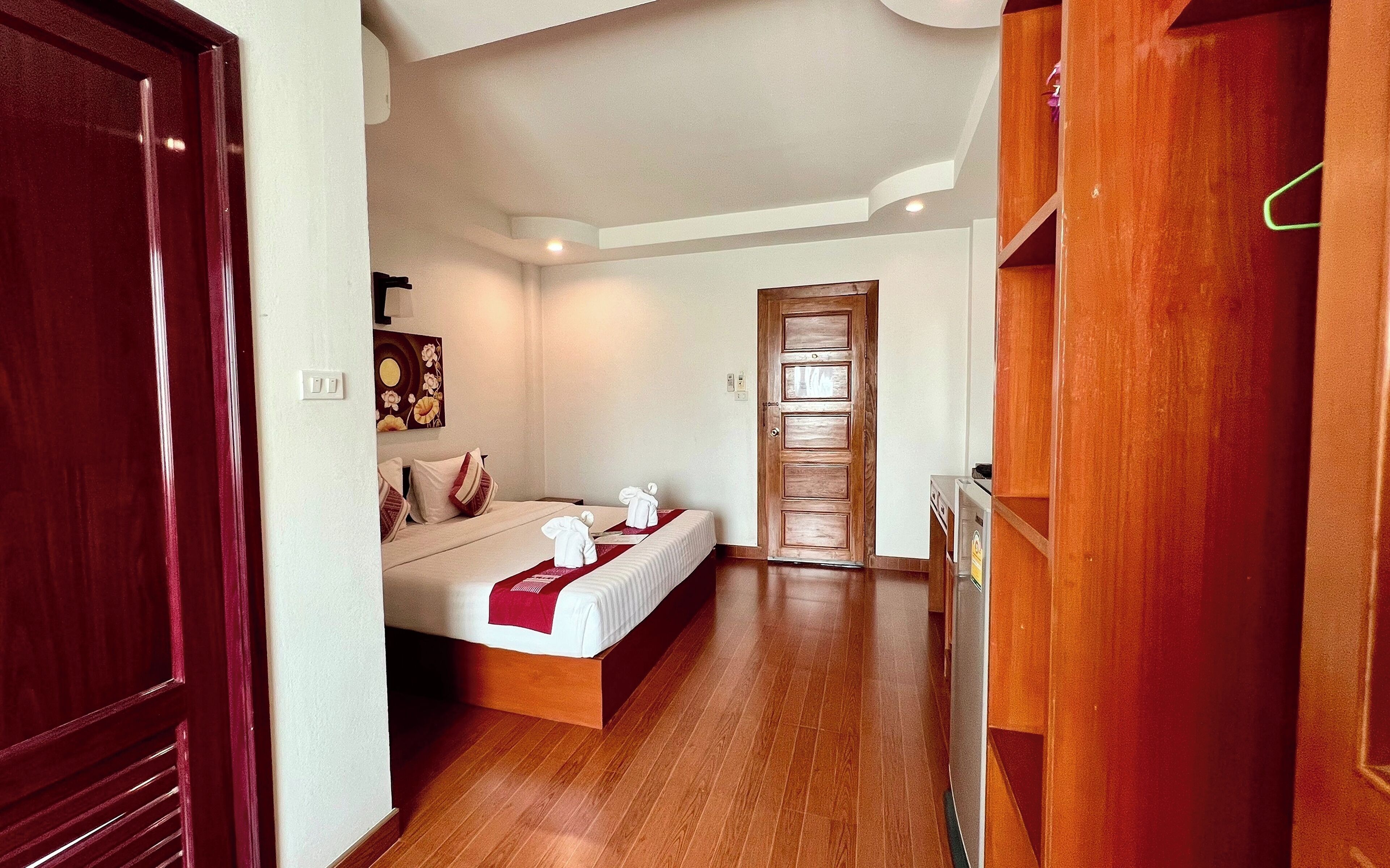 Quarto de hotel com piso de madeira, cama com roupa de cama branca e detalhe vermelho, guarda-roupa de madeira e porta que dá para outro quarto.