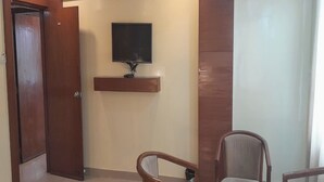 Quarto com porta de madeira, televisão montada na parede, uma prateleira pequena e duas cadeiras de madeira.
