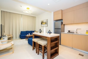 A modern living room with a blue sofa, a small dining table with stools, and a kitchen area with wooden cabinets and a refrigerator.
